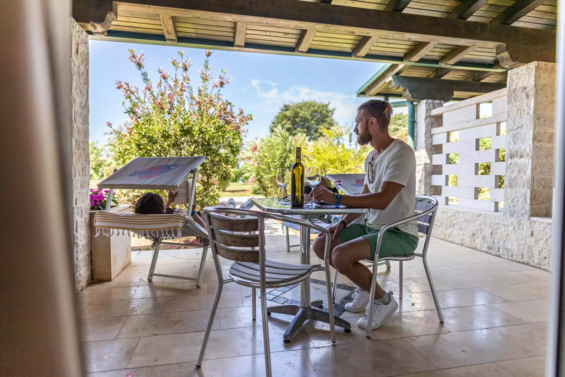 ragazzo con calice e bottiglia di vino al tavolo in veranda della villa nel campeggio mentre la ragazza legge un libro seduta sullo sdraio